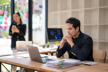 A man is sitting at a desk with a laptop and a stack of papers