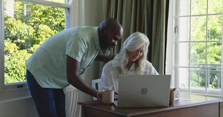 Diverse couple at home office, man arriving, pointing at laptop, moving mug to guide partner - Powered by Adobe