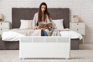Young woman with plaid reading book near electric convector heater in bedroom
