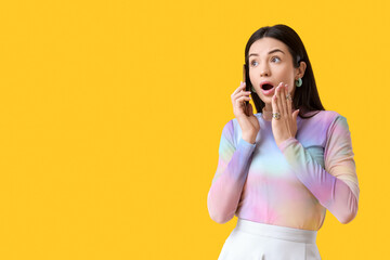 Shocked young woman talking by mobile phone on yellow background