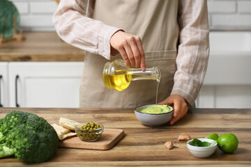 Woman pouring oil from jug into bowl with tasty peas hummus at table in kitchen