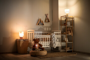 Interior of children's bedroom with baby crib, shelving unit, toys and glowing lamps at evening