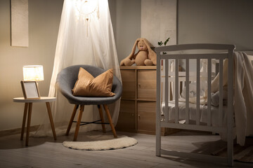 Interior of children's bedroom with baby crib, armchair, glowing lamps and toy bunny on commode at...
