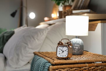 Alarm clock with glowing lamp on wicker table near bed in bedroom at night