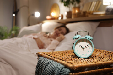 Alarm clock on wicker table against young woman sleeping under blanket in bedroom at night