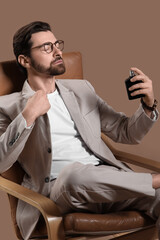 Handsome young man in light suit and eyeglasses with perfume bottle sitting on armchair against...