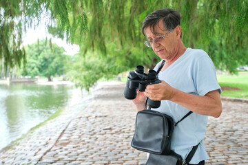 Senior Hispanic man taking out binoculars to watch birds in an urban park with a lake and trees that are home to this type of wildlife. Composition with copy space.