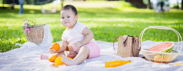Family enjoys leisure picnic in park with baby sitting on blanket holding orange surrounded by picnic baskets and fresh fruit in peaceful outdoor concept