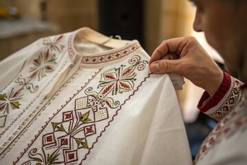 Close-up detail shot of intricate embroidery on traditional clothing, hands holding fabric with respect and care. Focus on texture and craftsmanship.