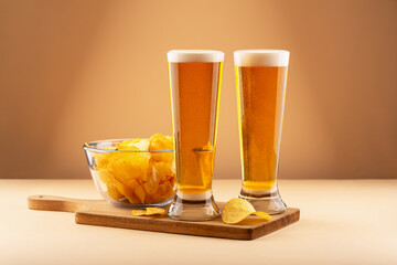 Crispy potato chips in glass bowl with two cold beers on wooden tray against warm background. Concept of bar snack, social drink and casual beer pairing. Beer and chips reflect comfort and taste