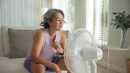 Asia adult people middle age old woman sit at sofa home feel hot flash fever use fan waving hand...