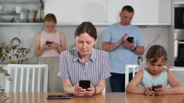 Two teenage girls sitting with parents at table in kitchen, engrossed in smartphones, oblivious to surroundings. Concept of family phubbing and addiction to mobile gadgets
