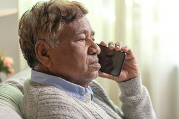 Closeup of sad elderly Asian man using mobile phone while sitting on sofa at home.