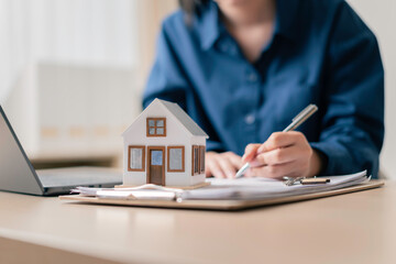homebuyer signing property purchase agreement at desk with laptop and miniature house model, finalizing mortgage and closing transaction