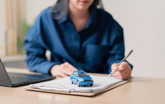 individual signing auto loan documents at a desk with a laptop and miniature blue car, illustrating digital financing and vehicle ownership