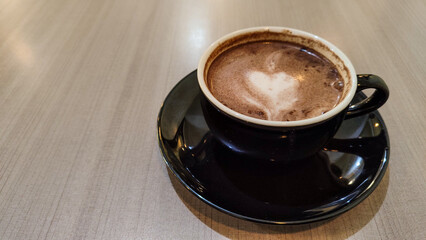 Top view of a cup of hot chocolate with heart-shaped latte art on a wooden table. Cozy and warm beverage in a black ceramic cup and saucer.