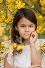 Little girl in white dress standing near blooming yellow trees and looking at camera, children portrait and childhood memories concept