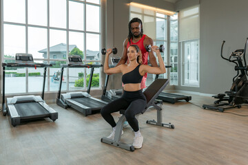 A woman perform a shoulder press with dumbbell while seated on a workout bench. Her trainer guide her, providing support and encouragement in a well-lit gym setting.