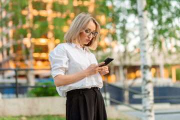 Young woman in glasses and white shirt and black trousers looking at phone against background of trees and building illuminated by sunset light.