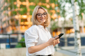 Young woman in white shirt with rolled up sleeves, wearing glasses, smiling, holding smartphone, against background of city street with trees and soft light.