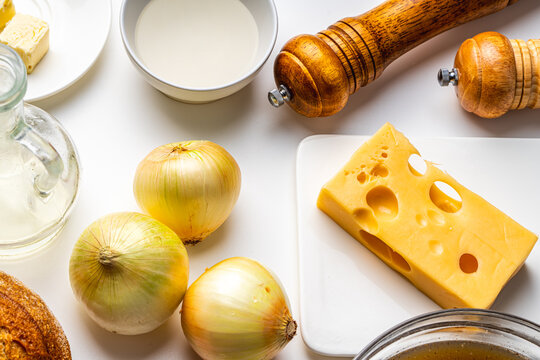 Ingredients for French onion soup. On the white table there is cheese, onions, bread, broth, mills with salt and pepper. Top view.