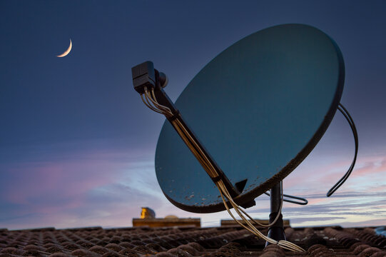 Satellite dish on a house roof with a crescent moon in the evening sky
