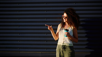 woman with sunglasses holding coffee cup pointing, casual outdoor style with bright smile. natural...