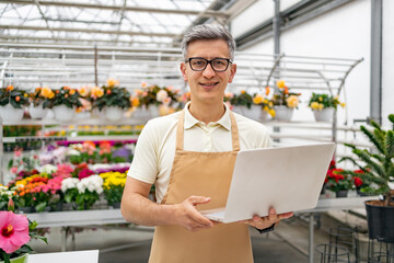 A florist, holding a laptop, smiles from within his vibrant flower shop, offering a welcoming atmosphere.