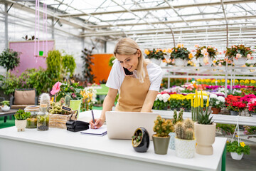 A Caucasian woman works at a florist shop, taking orders over the phone and using a laptop to manage sales while surrounded by various flowers and plants.