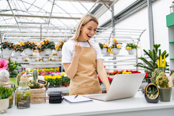 A cheerful Caucasian woman celebrates a successful sale in her flower shop, working at the table, in front of flowers and plants.