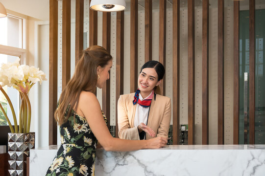 Friendly hotel staff in blazer with striped scarf warmly welcomes guest in sunflower dress at marble reception counter for room accommodation check in