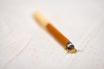 Close-up of cigarette resting against white fabric, leaving burn mark. Soft lighting highlights damage, contrasting with brown and white tones. Shallow depth of field focuses on burn area