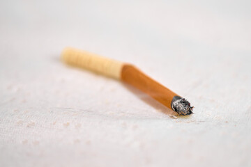 Single extinguished cigarette rests on light, textured surface. Orange filter contrasts with ash at tip. Soft lighting creates calm yet somber mood