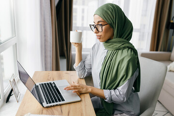 Side view of modern arabic businesswoman checking e-mail or corporate chat on laptop sitting at table at home with coffee mug in had, scrolling,typing on keyboard, working remotely