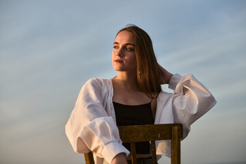Young woman in white blouse and black top sits on wooden chair by ocean. Golden hour light creates...