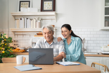 Asian senior man working with laptop at desk by supporting from cheerful wife at home. Couple spending time together.