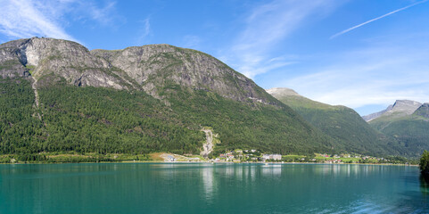 Panoramic view of Loen village, Norway, with the Innvikfjorden fjord in the foreground and majestic mountains and Hoven peak behind under a sunny sky, a typical northern fjord landscape