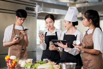 Asian female chef holding a tablet while her students looking at seasonal jars over kitchen counter.