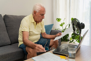 Senior man managing home finances and electricity bill on laptop during summer heat at home