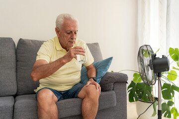 Senior man drinking water on sofa with electric fan cooling him down during summer heatwave at home