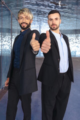 Vertical Portrait Two young businessmen in formal suits smiling confidently at the camera, giving thumbs up gesture inside a modern glass office with reflections and stylish interior design elements. 