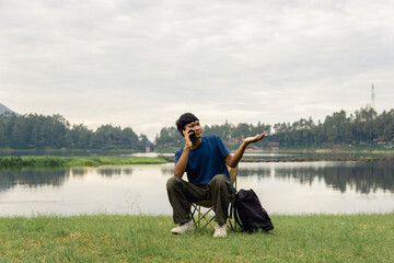 A young man sitting on a chair at lake with relaxing mood,  smiling and engaging in cheerful phone conversation.