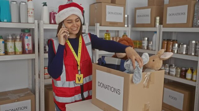 Woman in red vest and santa hat volunteering at charity donation center sorting toys and speaking on the phone in an indoor room filled with boxes and supplies.