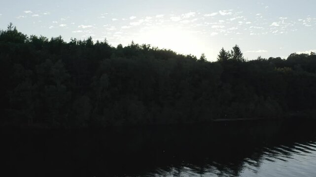 Aerial view of dark lake at sunset, sun low on horizon behind silhouetted forest, rippling water, Marcillac La Croisille, France