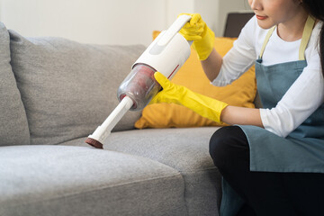 Asian female homeowner maid tidy up at home or apartment vacuuming dust and cleaning dirt from sofa in living room to perform household chores, housekeeping routine job using vacuum cleaner