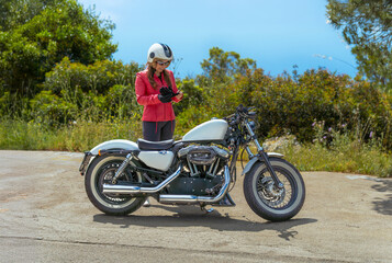 A beautiful young woman wearing a red jacket and open-face helmet putting on her gloves next to her customized Harley-Davidson motorcycle on a scenic coastal road.
