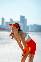 Female asian young jogger resting on beach in bright athletic wear with cityscape