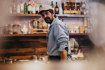 A man wearing a cap and denim shirt standing in a cozy kitchen ambiance filled with shelves of jars, bottles, and decorative elements.