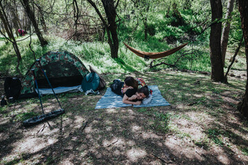 A pair enjoys time outdoors in a serene forest environment with a camping setup including a tent and hammock.