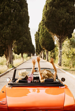 Two young women raising arms while driving orange convertible car on a country road surrounded by cypress trees
